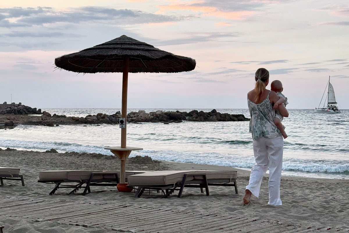 A woman in white clothing holds a baby on a sandy beach next to a thatched beach umbrella, with the ocean, rocky breakwater, and a sailboat visible in the background under a soft sunset sky.